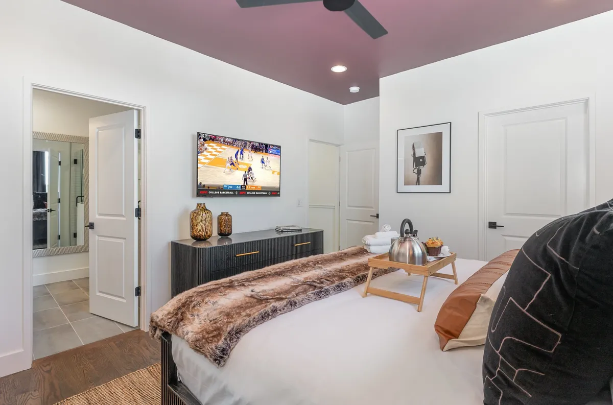 A cozy bedroom featuring a plush bed adorned with decorative throws, a television mounted on the wall displaying a basketball game, and a dresser with tasteful decor. The space exudes comfort and style, inviting guests to relax and unwind after a day in Nashville.