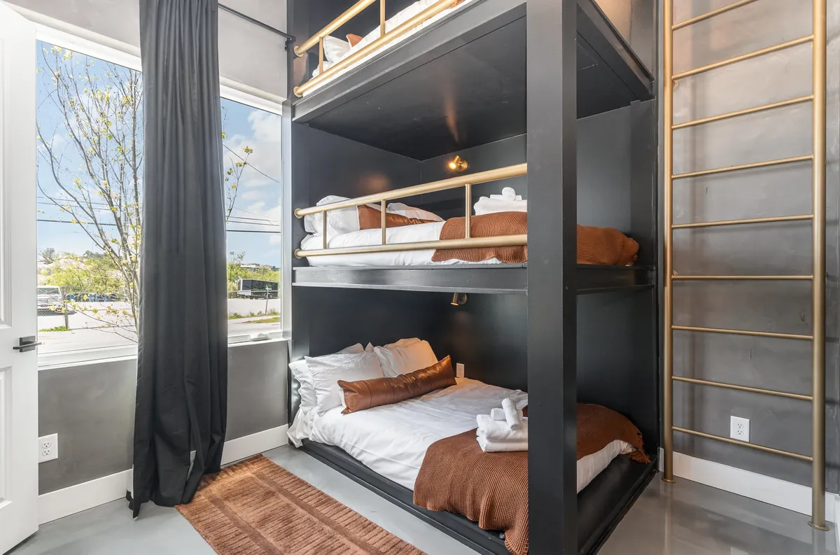 A spacious bunk room with sturdy bunk beds, a large window that offers plenty of natural light, a golden ladder, and a textured rug. The playful and inviting setting is perfect for kids or groups of friends to relax, socialize, or enjoy a late-night chat.