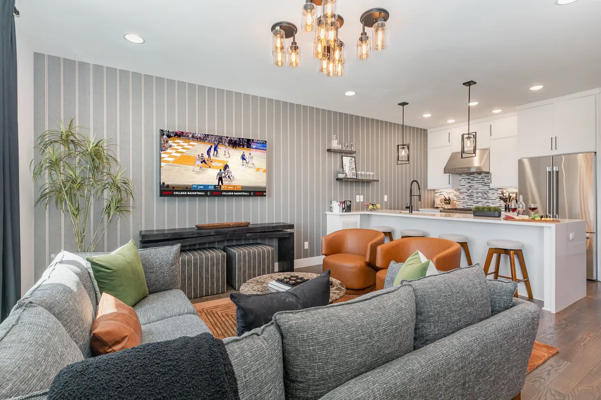 A spacious, inviting living room with a plush sectional sofa, central coffee table, kitchen island, and a large television. Modern lighting fixtures and an energetic layout set the stage for socializing, watching sports, or entertaining guests.