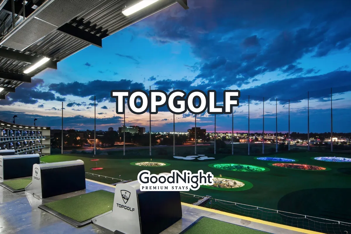 The image captures a lively scene at a modern golfing venue with multiple golfing bays, vibrant targets set against the ground, and the city skyline in the background. People are visible enjoying a game of golf, socializing, and dining, all while the sun sets, casting a warm, energetic glow over the space.