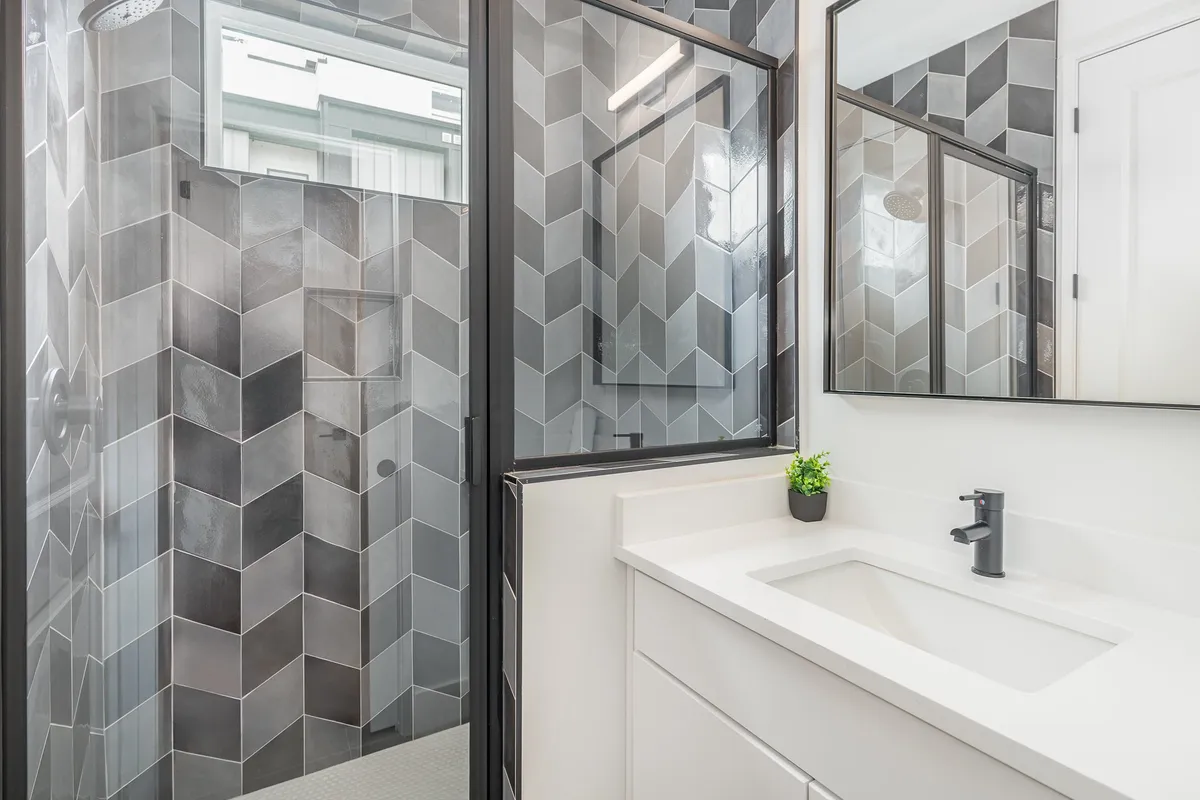 A modern bathroom featuring a glass-enclosed shower with chevron-patterned tiles, a sleek sink and faucet, and a large mirror. The space radiates cleanliness and contemporary style, ideal for spa-inspired self-care rituals.